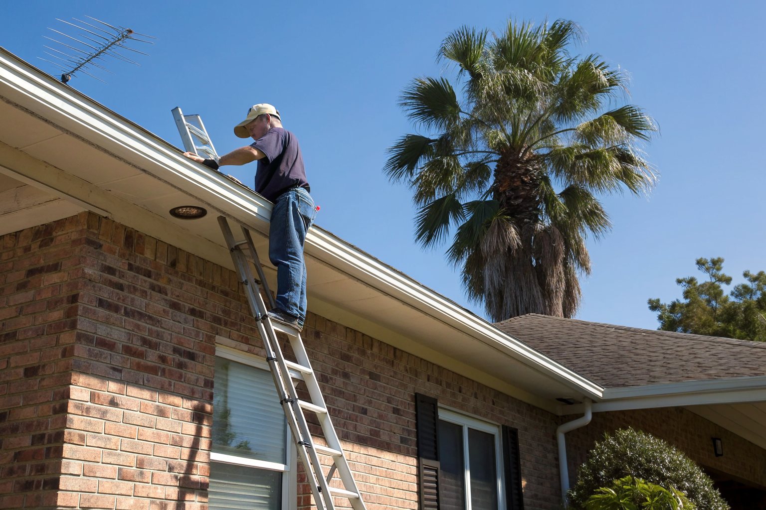 A roofer on a ladder checking the shingles and gutter edge of a brick home